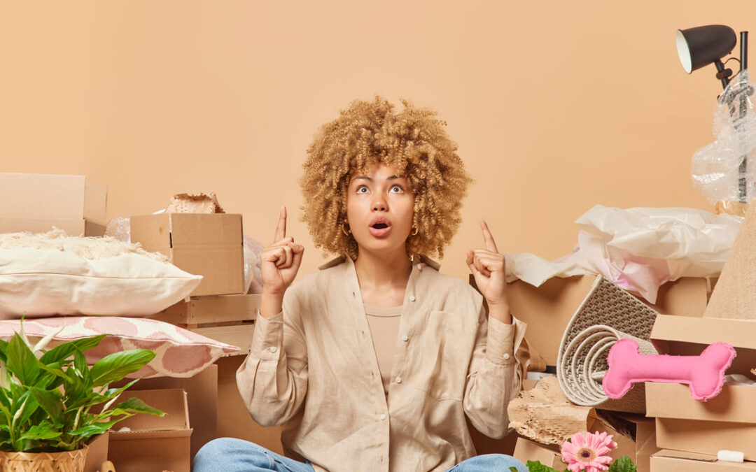 Shocked curly haired adult woman points overhead hold breath from amazement surrounded by carton